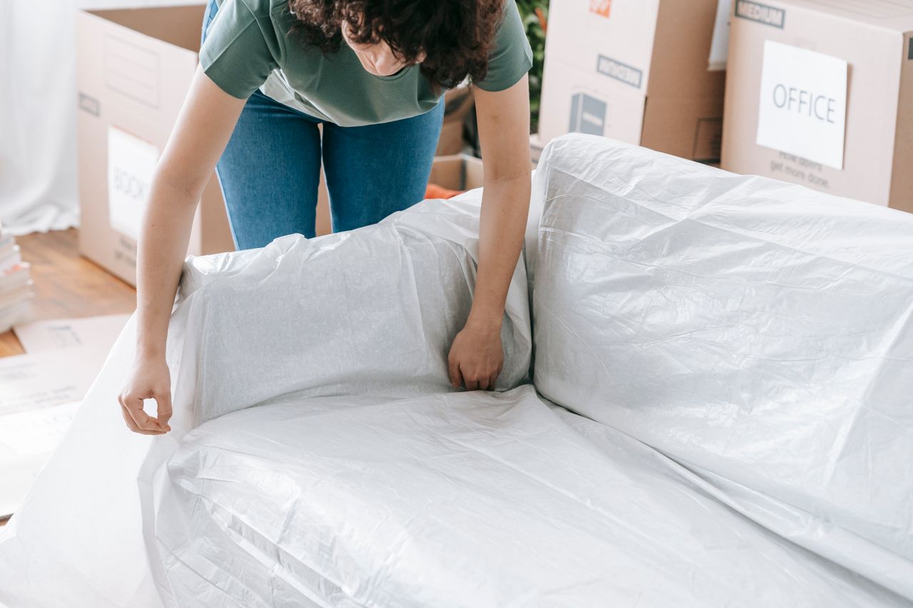 A woman thoughtfully covers a sofa with protective wrapping while boxing items in a well-lit room.